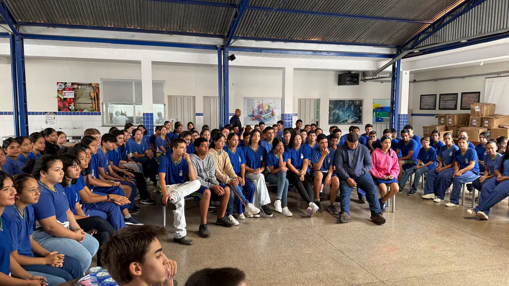 Alunos de uniforme azul sentados no pátio coberto da escola acompanham a roda de conversa