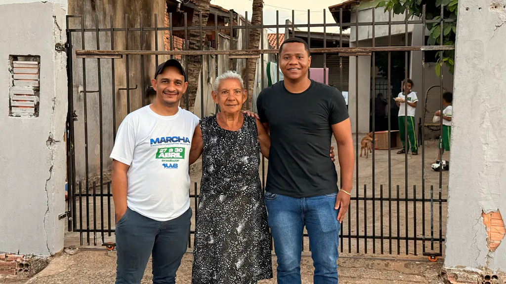 Pelézinho, de camiseta branca, Dona Benvina e Nunes, presidente do Rotary, sorrindo em frente ao portão da casa dela em Nova Mutum