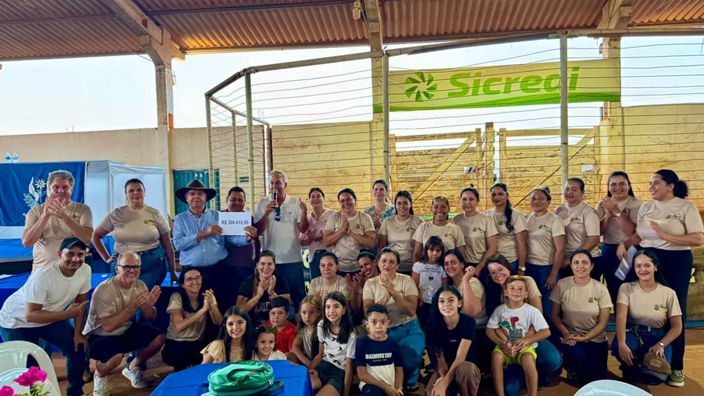 Equipe e voluntários do leilão da APAE de São José do Rio Claro reunidos no recinto, aplaudindo, enquanto dois homens seguram a placa com o valor arrecadado de R$ 306.410,00