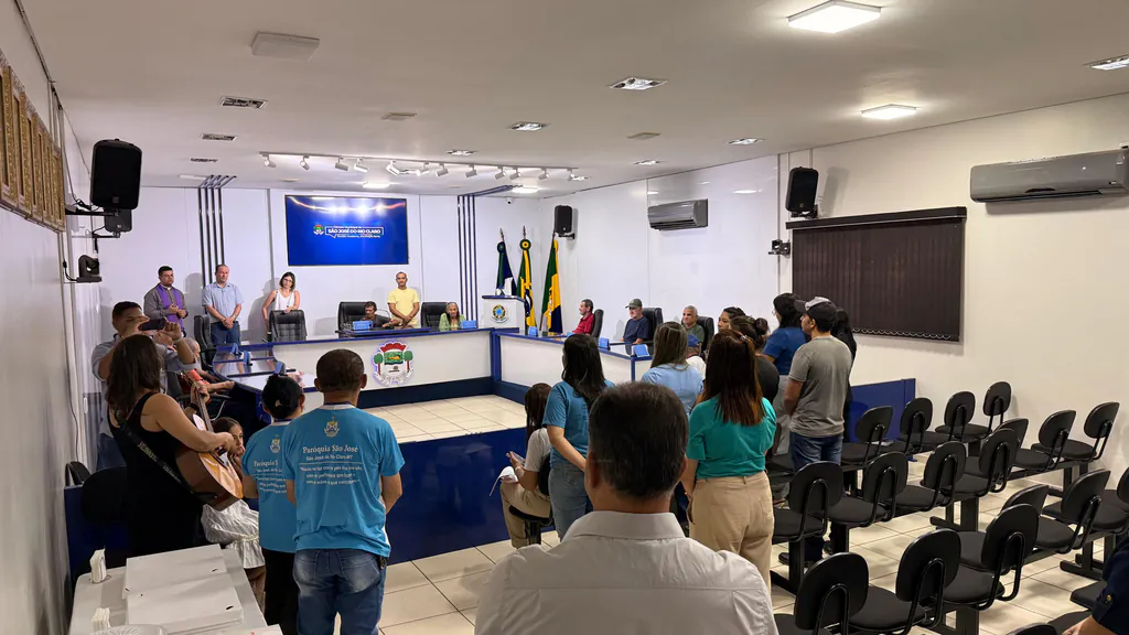 Convidados de pé no plenário da Câmara durante o momento de oração, com idosos sentados na bancada ao fundo