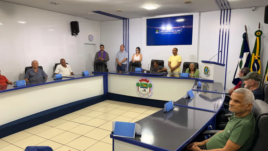 Idosos sentados nas cadeiras da bancada do plenário da Câmara Municipal de São José do Rio Claro, com o padre e convidados de pé ao fundo